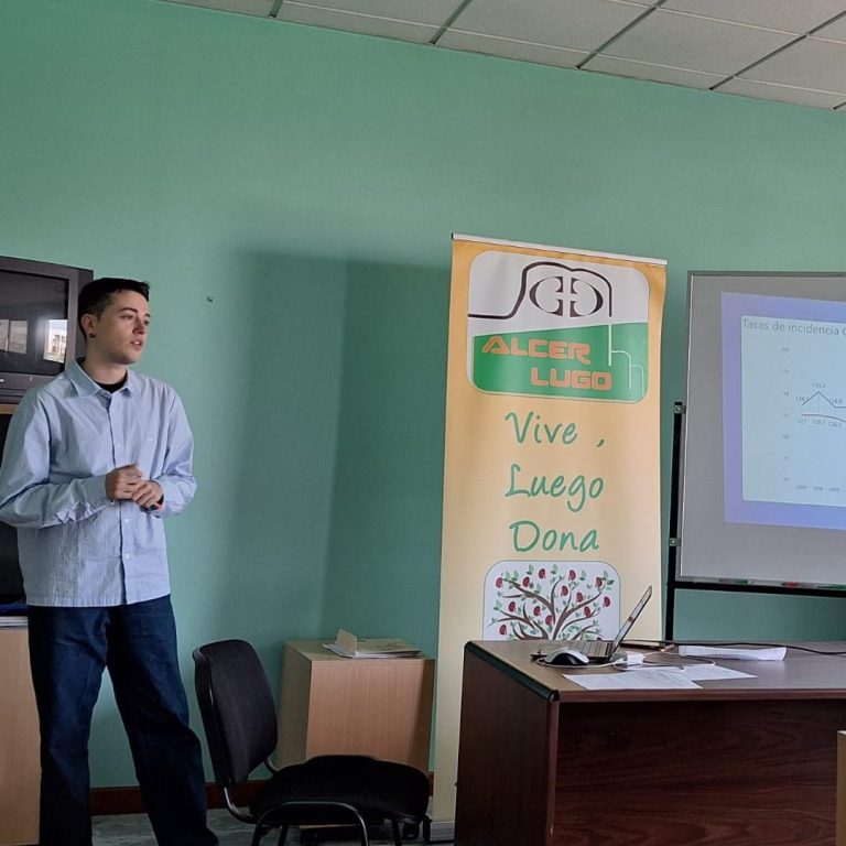 Hombre presenta en una sala con pizarra y banner de evento educativo.